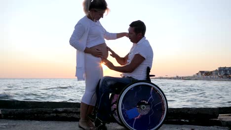 invalid-spouse-in-wheelchair-with-pregnant-female-at-sunset,-handicapped-man-in-wheelchair-caresses-woman