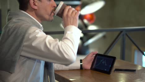 Mature-handsome-man-drinks-coffee-and-monitors-cryptocurrency-trading-on-tablet-in-a-cafe