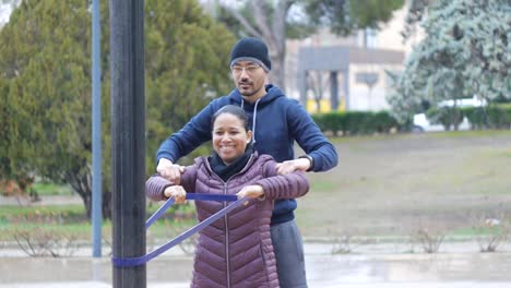 Al-aire-libre-del-entrenamiento-de-pareja.-Pareja-joven-de-América-Latina-hace-fila-de-resistencia-banda-juntos-en-el-Parque-otoño-como-parte-del-programa-habitual-de-ejercicios.-Hombre-con-lentes-ayuda-a-risa-mujer-para-tirar.