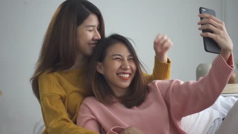 Beautiful-young-asian-women-LGBT-lesbian-happy-couple-sitting-on-sofa-hug-and-using-phone-taking-selfie-together-bedroom-at-home.-LGBT-lesbian-couple-together-indoors-concept.-Spending-nice-time-home.