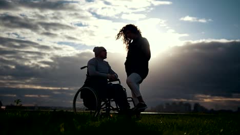 Cuidado-de-personas-con-discapacidad---hombre-en-silla-de-ruedas-con-mujer-hablando-en-la-puesta-de-sol-nublado