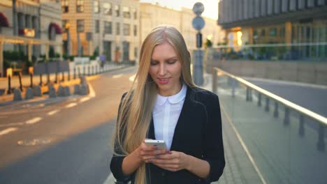 Formellen-Business-Frau-gehen-auf-der-Straße.-Elegante-blonde-Frau-im-Anzug-und-gehen-auf-der-Straße-und-Surfen-Smartphone-mit-Lächeln-städtischen-Hintergrund