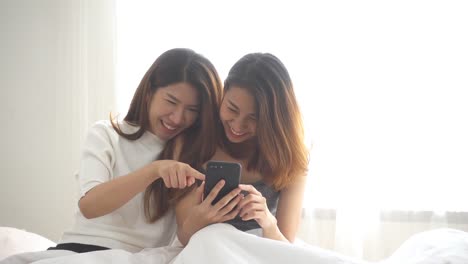 Slow-Motion---schöne-junge-asiatische-Frauen-LGBT-lesbische-glückliche-Paar-sitzt-am-Bett-Umarmung-und-Telefon-zusammen-Schlafzimmer-zu-Hause-verwenden.-Lesbisches-Paar-gemeinsam-im-Haus-Konzept.-Schöne-Zeit-zu-Hause-verbringen.