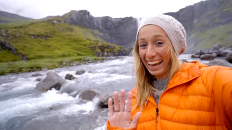 Young-woman-taking-selfie-portrait-with-magnificent-waterfall-in-Iceland,-blowing-a-kiss-to-camera.-People-travel-exploration-concept