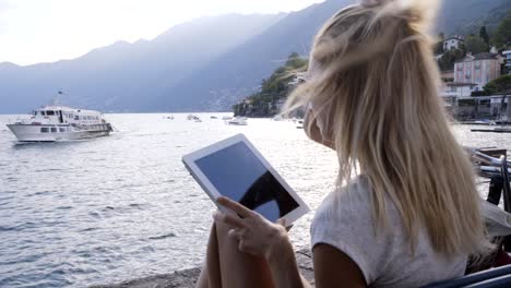 Young-woman-sitting-on-a-bench-in-a-town-facing-the-lake-and-mountains-using-a-digital-tablet-technology--Mobile-apps,-online-reservation-concept