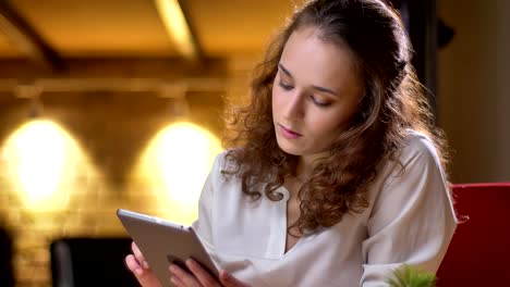 Portrait-of-young-curly-haired-businesswoman-seriously-watching-into-tablet-and-raising-her-eyes-to-camera-in-office.