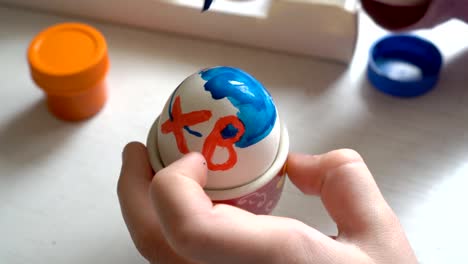 Close-up,-the-child-paints-a-white-Easter-egg-with-a-brush-and-blue-paint.-The-view-from-the-top.-4K.