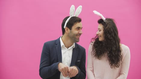 Beautiful-young-couple-standing-on-pink-background.-At-the-same-time,-they-smile-to-recreate-the-movements-of-hares-after-kissing.-Together-with-the-pink-paws-on-the-head.-Happy-family-is-preparing-for-Easter.