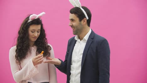 Young-couple-are-beautiful-on-pink-background.-During-this-time,-they-are-dressed-in-rabble-ears.-Looking-at-each-other.-A-man-shows-carrots-and-his-wife-starts-to-laugh.-A-young-man-begins-to-choke-the-girl.