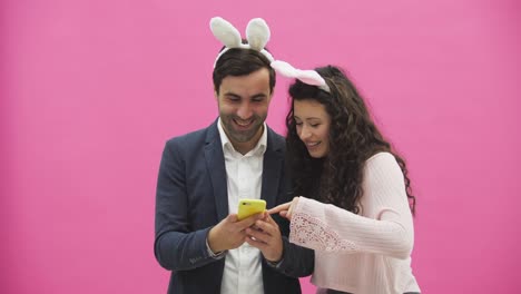 Junge-Liebhaber-Paar-auf-rosa-Hintergrund.-Mit-reißenden-Ohren-auf-dem-Kopf.-Während-dieses-Osterfotos,-sephi-tun-am-Telefon-und-schauen-sie-freuen.