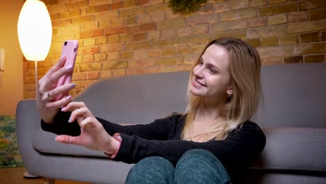 Retrato-en-primer-plano-de-la-joven-estudiante-tomando-selfies-en-el-teléfono-y-sonriendo-mientras-se-sienta-en-el-suelo-apoyado-en-el-sofá-en-el-interior-acogedor-hogar