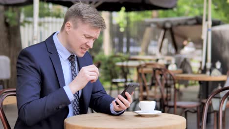 Businessman-Upset-by-Loss-on-Smartphone,-Sitting-in-Outdoor-Cafe