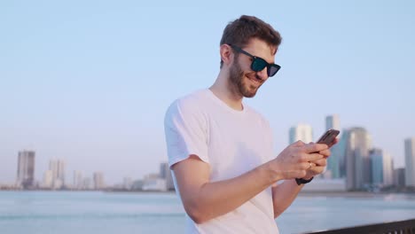 Attraktiver-und-hübscher-junger-Mann-mit-schönem-Lächeln-und-Hipster-FelMillennial-Generation-z-sieht-aus,-scrollt-durch-Social-Media-Feeds-auf-Smartphone,-kichert-und-lächelt-von-Nachrichten-über-Anwendungen