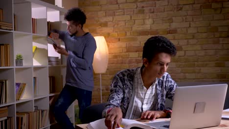 Closeup-shoot-of-two-culturally-diverse-students-learning-in-the-school-library-indoors.-Indian-male-studying-online-on-the-laptop-while-african-american-man-using-the-tablet-on-the-background