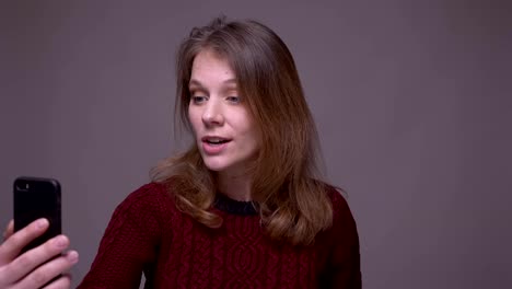 Young-female-student-talking-in-videochat-on-smartphone-on-gray-background.