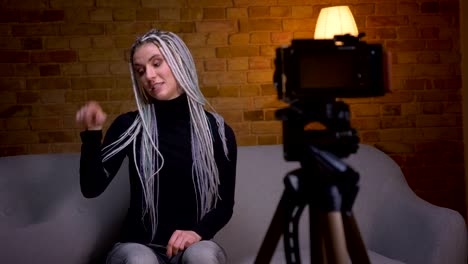 Closeup-shoot-of-young-attractive-female-blogger-with-dreadlocks-streaming-live-talking-on-camera-sitting-on-the-couch-indoors