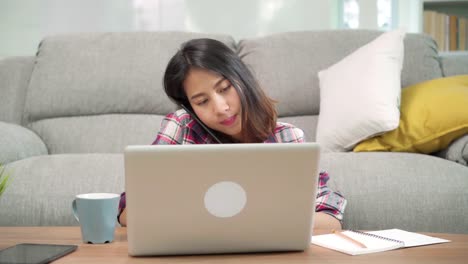 Freelance-Asian-woman-working-at-home,-business-female-working-on-laptop-and-using-mobile-phone-talking-with-customer-on-sofa-in-living-room-at-home.-Lifestyle-women-working-at-home-concept.