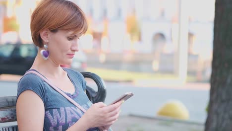 Woman-Using-Smartphone-Relaxes-on-the-Bench-t-the-street.-SLOW-MOTION,-4K.-Young-Millennial-Woman-in-Arboretum-making-gestures-on-Phone-Display.