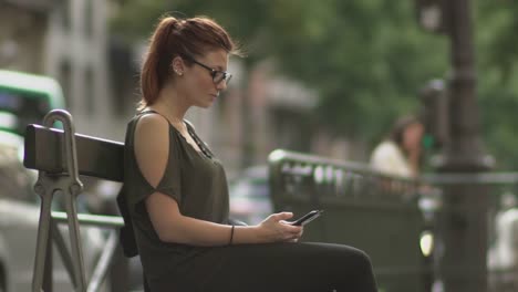 Attractive-redhead-woman-with-glasses,-freckles,-piercings-and-red-hair-writing-a-text-message-on-her-smartphone-sitting-on-street-bench,-during-sunny-summer-in-Paris.-Slow-motion.-Trendy.