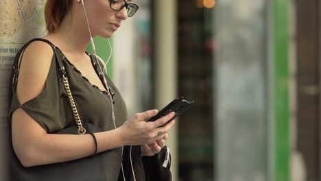 Atractiva-mujer-caucásica-con-auriculares,-gafas,-pecas,-piercings-y-pelo-rojo-escuchando-música-y-escribiendo-mensajes-de-texto-mientras-espera-en-la-parada-de-autobús-en-París.-A-cámara-lenta.-Clima-soleado.