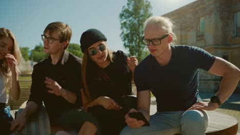 Stilvolle-coole-Teenager-Chillen-auf-den-Bänken-im-Park,-mit-Smartphone.-Zwei-Mädchen-und-zwei-Jungen-im-Gespräch-und-Spaß-in-den-sozialen-Medien.-Hip-Park-in-Gentrified-Part-of-City