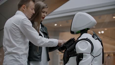 Ein-Mann-und-eine-Frau-in-der-Mall-interagieren-mit-einem-Roboterberater,-indem-sie-auf-den-Bildschirm-tippen-und-lächeln.-Cyborg-Android-hilft-Menschen.