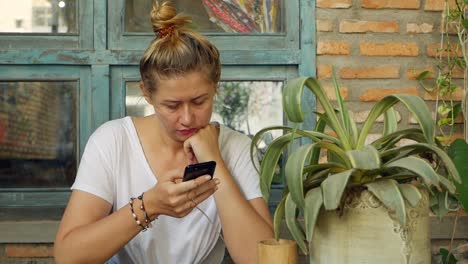 Pensive-girl-uses-a-smartphone,-writes-a-message,-chatting.-Woman-looking-for-information-in-the-phone-and-sad.-Sitting-in-restaurant-alone.-Breakfast-time.-Technology-and-entertainment-concept