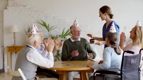 Tracking-shot-of-nurse-bringing-birthday-cake-for-elderly-man-playing-cards-with-aged-friends-sitting-at-table-in-nursing-home.-Happy-senior-man-getting-wishes,-kisses-and-hugs-and-blowing-candle-out