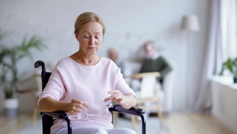 Behinderte-ältere-Frau-sitzt-im-Rollstuhl-und-schaut-nachdenklich-weg.-Traurige-Patientin-schaut-in-die-Kamera,-wenn-junge-Krankenschwester-bringt-ihre-Vitaminpillen-und-Glas-Wasser