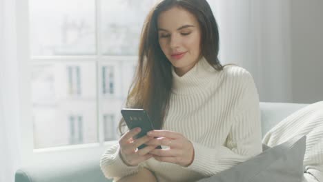 Hermosa-mujer-joven-usando-Smartphone-sonrientemente,-mientras-está-sentada-en-la-silla.-Sensual-chica-usando-suéter,-Surfs-Internet,-publicaciones-en-las-redes-sociales-mientras-se-relaja-en-el-apartamento-acogedor.-Retrato-de-primer-plano