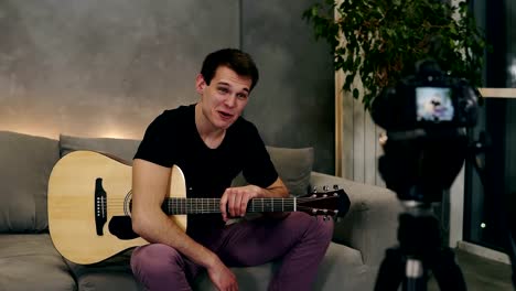 Handsome-caucasian-man-in-black-T-shirt-making-video-blog-about-musical-instruments-or-recording-the-song-for-his-followers-while-sitting-on-a-couch-at-home.-Recording-camera-on-a-foreground