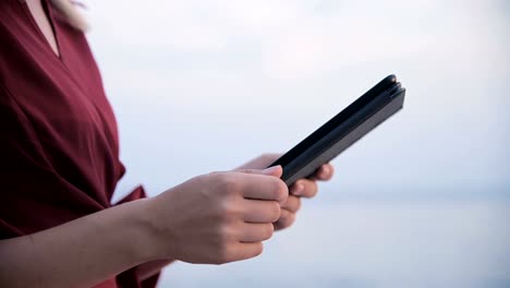 Close-up-An-attractive-young-girl-hands-in-a-summer-red-dress-sits-on-a-stone-by-the-sea-in-the-evening-and-looks-at-something-on-a-tablet.-Swipe-across-the-screen