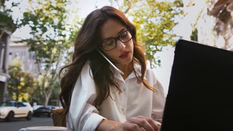 Mujer-de-negocios-con-gafas,-camisa-blanca.-Sentado-en-la-mesa-con-taza-de-café-y-portátil-en-una-cafetería-al-aire-libre.-Hablando-por-teléfono-celular,-escribiendo.-Cámara-lenta