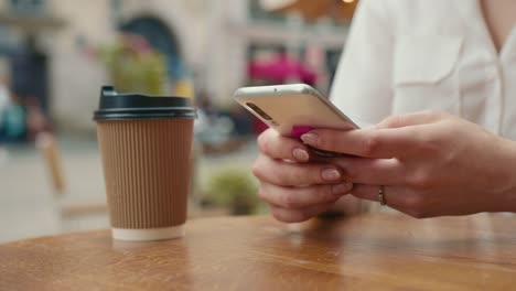 Disparo-de-primer-plano.-La-chica-está-enviando-mensajes-de-texto-en-su-teléfono-inteligente.-Hay-una-taza-de-café-en-la-mesa.-Está-sentada-en-un-café.-4K