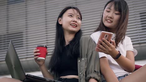 Beautiful-happy-smiling-asian-girls-sitting-near-big-urban-building-and-using-phone-to-watch-photo