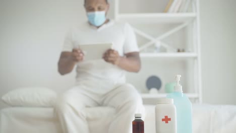 Close-up-of-sanitizer-and-pills-with-blurred-African-American-man-using-tablet-at-the-background.-Ill-male-patient-in-face-mask-sitting-on-bed-in-hospital-ward-surfing-Internet.-Covid-19-pandemic.