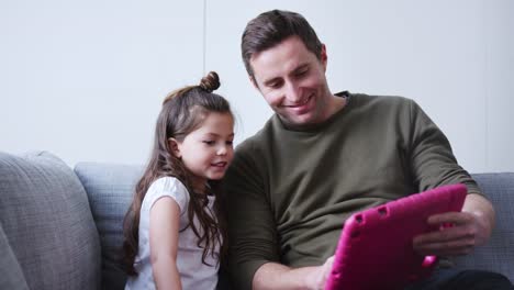 Vater-und-Tochter-sitzen-auf-Sofa-zu-Hause-spielen-zusammen-auf-digitalen-Tablet-in-rosa-Fall-zu-Hause