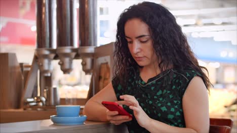 La-chica-está-escribiendo-en-el-teléfono-inteligente-mientras-está-sentada-en-una-cafetería.-Retrato-de-cerca-de-una-joven-feliz,-chica-sentada-en-un-café-bebiendo-café-por-la-mañana-en-la-ciudad.