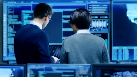 In-the-System-Control-Room-IT-Specialist-and-Project-Engineer-Have-Discussion,-they're-surrounded-by-Multiple-Monitors-with-Graphics.-They-Work-in-a-Data-Center-on-Data-Mining,-AI-and-Neural-Networking.