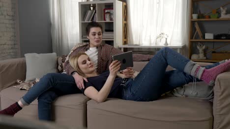 Lesbian-couple-is-resting-on-the-couch,-laughing-and-watching-funny-movie-on-tablet-60-fps