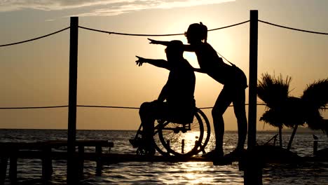 lucky-disabled-with-girlfriend-look-into-distance-and-shows-his-arms-to-horizon-on-background-sunset-at-over-ocean