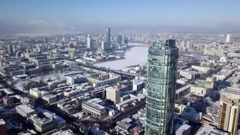 Aerial-view-of-skyscraper-is-in-the-middle-of-the-city-in-winter,-blue-sky-sky-and-snowy-roofs-of-buildings-background