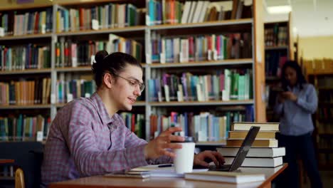 Estudiante-hombre-hipster-guapo-joven-está-sentado-en-mesa-en-luminoso-gran-biblioteca-con-el-portátil-delante-de-él,-la-impresión-y-tomando-café.-Él-está-sonriendo.