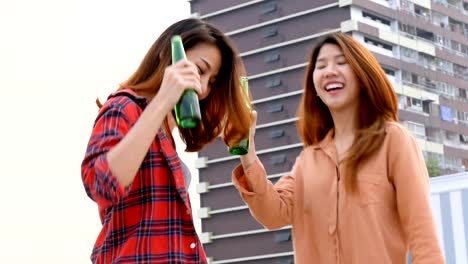 Mujer-asiática-joven-pareja-de-lesbianas-bailando-y-tintineantes-botellas-de-cerveza-fiesta-en-la-azotea.