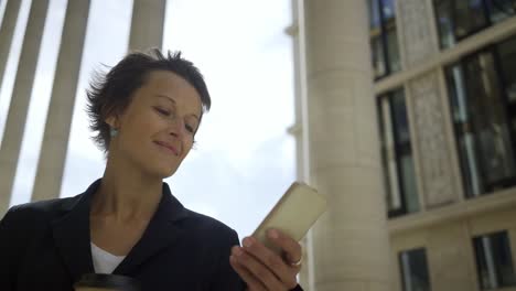 Panning-of-confident-businesswoman-with-short-hair-using-application-on-her-smartphone-outdoors,-takeaway-coffee-cup-in-her-hand