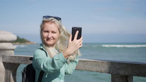 Junge-schöne-schlanke-Frau-mit-langen-blonden-Haaren-mit-Sonnenbrille-und-grünes-Hemd-stehen-in-der-Nähe-von-Palme-und-Selfie-auf-Handy-auf-blauem-Himmel-und-Meer-Hintergrund-zu-machen.-Mädchen-mit-smartphone