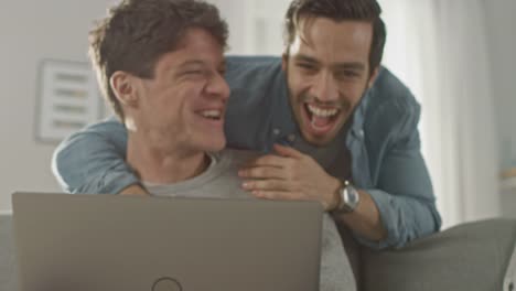 Entzückende-männliche-Gay-paar-verbringen-Zeit-zu-Hause.-Junger-Mann-arbeitet-auf-einem-Laptop,-seine-Partnerin-von-hinten-kommt-und-sanft-umarmt-ihn.-Sie-lachen-und-Hände-berühren.-Zimmer-ist-Modern-eingerichtet.