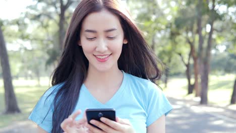 Hermosa-asiática-joven-con-smartphone-en-el-parque.-Mujer-con-smartphone-de-árbol-un-natural-en-el-fondo.