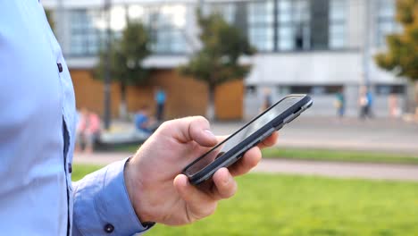 Close-up-hand-of-young-businessman-holding-and-touching-smartphone.-Unrecognizable-man-standing-on-urban-street-and-browsing-phone.-Guy-using-gadget-for-work.-Slow-motion-Side-view