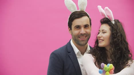 Young-creative-couple-on-pink-background.-With-hackneyed-ears-on-the-head.-During-this,-the-wife-holds-decorative-multicolored-eggs-in-her-hands.-Gently-kissing-her-husband-looking-at-the-camera-together.